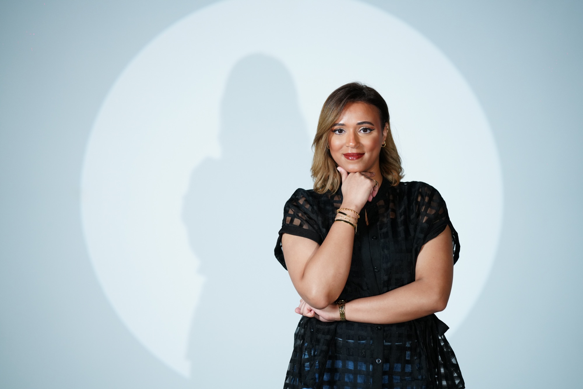 Zahra Amin smiling, resting her chin on her hand, in front of a spotlight on a pale blue wall